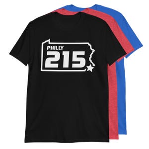 May include: Black t-shirt with a white outline of the state of Pennsylvania and the text "PHILLY 215" inside. The shirt is shown with red and blue t-shirts behind it.