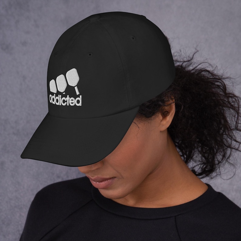 May include: Black baseball cap with white embroidery of three paddleball paddles and the word "addicted".
