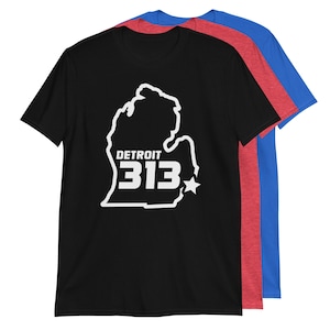 Puede incluir: Camiseta negra con un contorno blanco del estado de Michigan y el texto "DETROIT 313" dentro del contorno. La camiseta se muestra con camisetas rojas y azules detrás de ella.