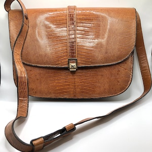 May include: A brown, textured leather crossbody bag with a flap closure. The bag features a long, adjustable strap and a decorative buckle on the front. The bag's design includes a reptile-like pattern, giving it a unique look.