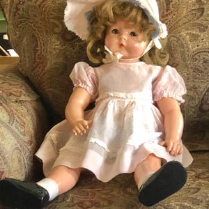 May include: A vintage doll with blonde hair, wearing a pink dress and a matching bonnet. The doll has a sweet expression and is seated in a patterned armchair. She is wearing black shoes and white socks.