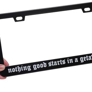 May include: Black metal license plate frame with the text "nothing good starts in a getaway car" in white lettering.