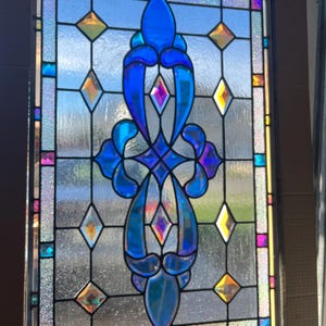 May include: A stained glass window with a central blue design and diamond-shaped accents in various colors. The window features a symmetrical pattern with a clear background and a black frame. The glass has a textured appearance.