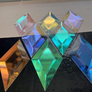 May include: Assortment of iridescent diamond-shaped glass tiles in various colors, including blue, green, gold, and purple. The tiles are arranged in a starburst pattern on a black surface.