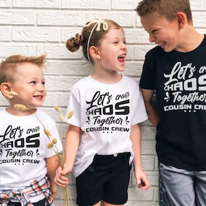 May include: Three children wearing white and black t-shirts with the text "Let's create chaos together cousin crew". The children are standing in front of a white brick wall.