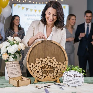 May include: A wooden guest book with a tree design, surrounded by small wooden leaf-shaped pieces. The guest book is on a table with a green cloth, with a sign that says "Guest Book". People are in the background.