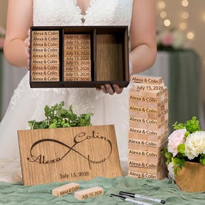 May include: A bride holds a wooden box with engraved blocks, a wooden sign with an infinity symbol, and a stack of blocks. The sign and blocks have names and a date. The table is decorated with flowers and greenery.