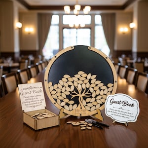 May include: A wooden guest book alternative with a tree design, surrounded by leaf-shaped wooden pieces. Includes a box for the leaves and two signs that read "Guest Book". The guest book is on a long wooden table.