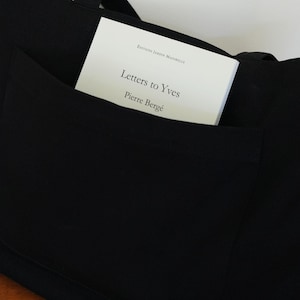 Op de afbeelding: Een zwarte tote bag met een groot buitenvak. In het vak zit een wit boek met zwarte tekst die luidt Editions Jardin Majorelle, Letters to Yves en Pierre Bergé. De tas heeft twee zwarte stoffen handvatten aan de bovenzijde.