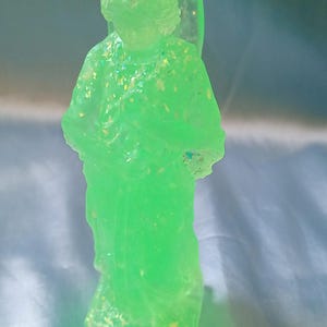 May include: A translucent green angel figurine with glitter embedded throughout. The angel is standing with its hands clasped in front. The wings are visible on the back. The figurine is on a light blue surface.