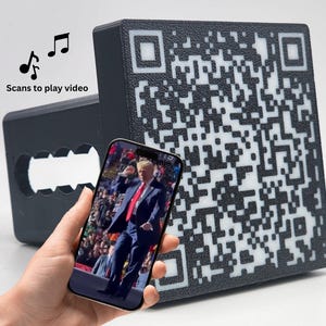 May include: A black QR code block and a black rectangular object with a cutout are displayed. A smartphone shows a video of a person in a suit. The text "Scans to play video" is above the objects.