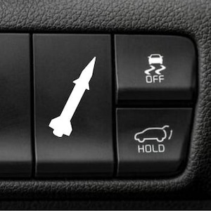 May include: A black plastic car dashboard button featuring a white decal of a missile. To the right, two additional buttons are visible: one with a car traction control icon labeled OFF and another with a car trunk icon labeled HOLD.