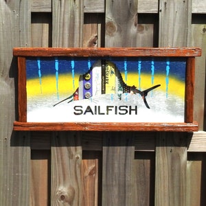 May include: A wooden framed artwork featuring a sailfish silhouette with a blue and yellow background. The sailfish is made of various beer can tabs and the artwork is titled "SAILFISH".