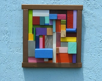 May include: A colorful abstract wall art piece made of wood blocks in various shapes and sizes. The blocks are painted in shades of red, blue, green, yellow, purple, and orange. The piece is framed in a brown wooden frame.