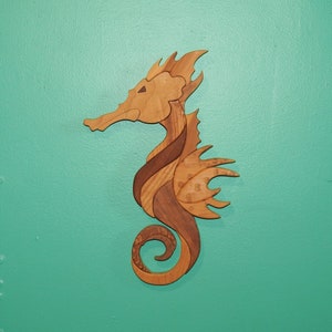 May include: Wooden seahorse wall art. The seahorse is crafted from various wood tones, including light and dark browns. The seahorse is mounted on a teal wall. The seahorse has a detailed design, with a curled tail and a distinctive head.