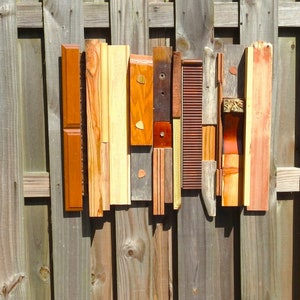May include: Abstract wooden wall art composed of various wood pieces in shades of brown, orange, and natural wood tones. The arrangement includes rectangular, square, and textured elements, some with decorative details. The artwork is mounted on a wooden fence.