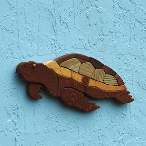 May include: A wooden turtle sculpture with a brown body and a shell made of different colored wood pieces. The turtle is swimming with its head and legs extended.