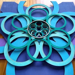 May include: A blue and green layered paper art piece with a circular design. The piece is made of multiple layers of paper, each with a different shade of blue and green. The center of the piece is a smaller circle with a series of concentric circles.