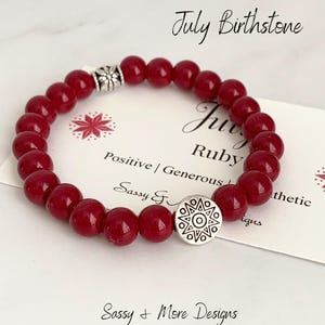 May include: Red gemstone bead bracelet with a silver charm featuring a starburst design. The bracelet is on a white card with the text "July Birthstone Ruby Positive | Generous Sassy & More Designs".