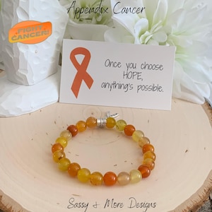 May include: Orange and yellow beaded bracelet with a silver charm that says "Hope". The bracelet is on a wooden surface with a card that says "Appendix Cancer" and "Once you choose HOPE, anything's possible."