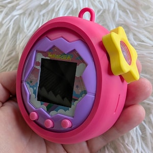 Puede incluir: Un juego electrónico portátil rosa con un borde de pantalla morado y azul y la palabra "Magagotchi" impresa en la pantalla. Tiene un botón amarillo en forma de estrella y tres botones rosas debajo de la pantalla. Un pequeño lazo rosa en la parte superior.