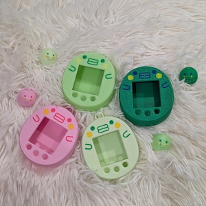 May include: Four colorful handheld electronic pet game devices in green, pink, and light green, with small matching character figures. The devices have a screen and buttons, and are arranged on a fluffy white surface.
