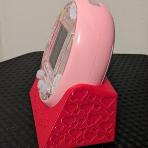 May include: A pink handheld electronic device sits in a red stand. The stand features a heart pattern. The device has a clear screen and buttons. The stand is designed to hold the device upright.