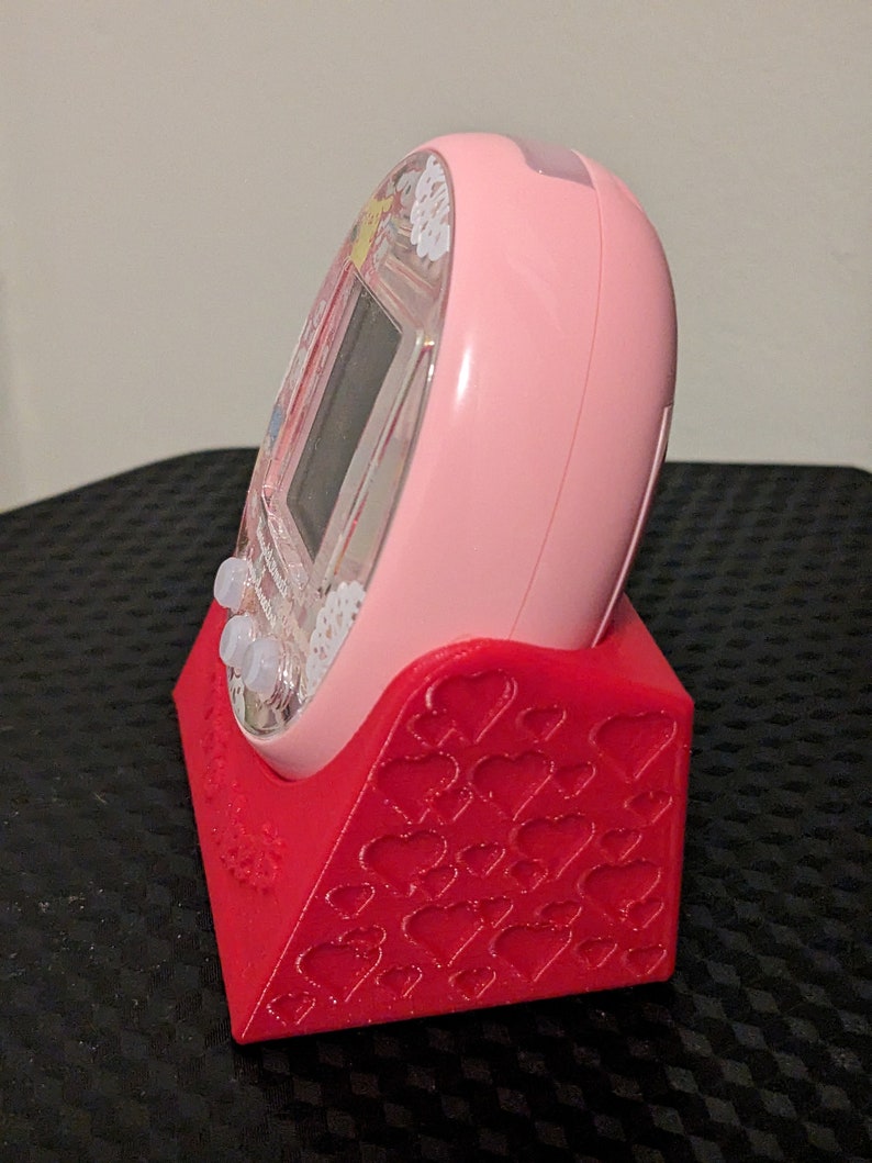 May include: A pink handheld electronic device sits in a red stand. The stand features a heart pattern. The device has a clear screen and buttons. The stand is designed to hold the device upright.
