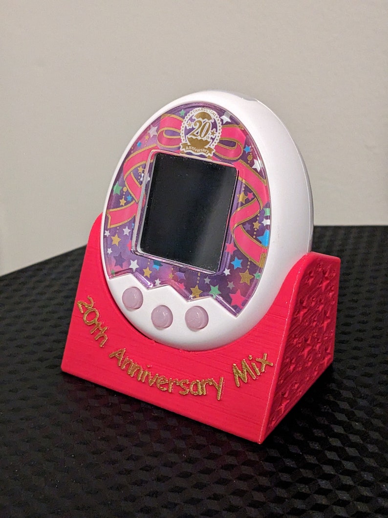 May include: A white and pink 20th Anniversary Mix digital pet game with a black screen, on a red stand. The stand has the text "20th Anniversary Mix" in gold lettering. The game features a ribbon and star design.