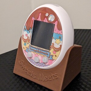 May include: A white digital pet device with a decorative faceplate featuring a chocolate and dessert theme. The device is in a brown stand with the words "Sweets Meets" engraved on it. The screen is black.