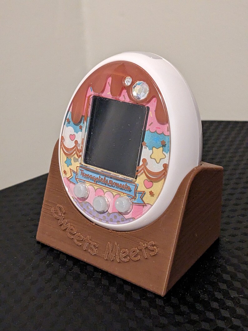 May include: A white digital pet device with a decorative faceplate featuring a chocolate and dessert theme. The device is in a brown stand with the words "Sweets Meets" engraved on it. The screen is black.