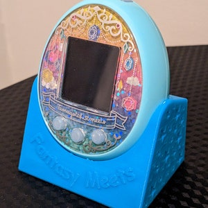 May include: A light blue Tamagotchi Meets digital pet device, on a matching blue stand. The device has a colourful, glittery display with the text "Tamagotchi Meets" and "Fantasy Meets". The stand has the words "Fantasy Meets" on it.