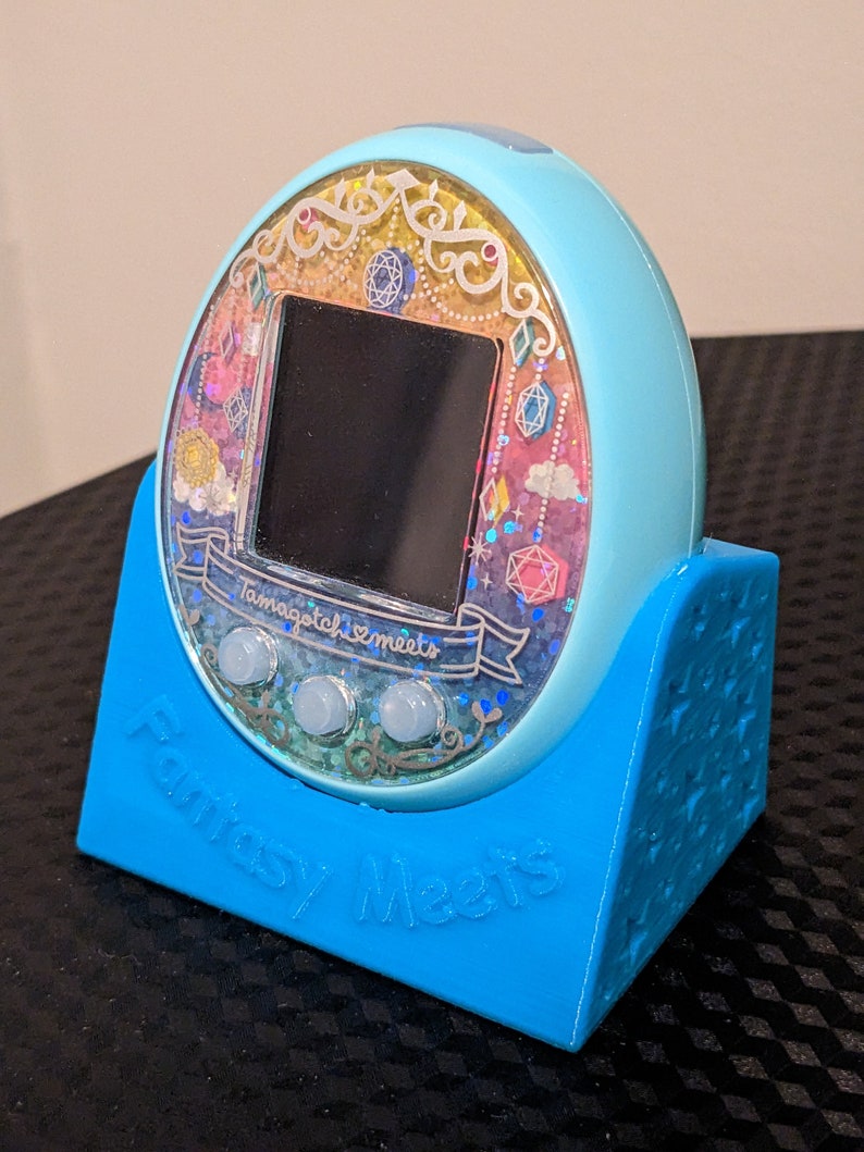 May include: A light blue Tamagotchi Meets digital pet device, on a matching blue stand. The device has a colourful, glittery display with the text "Tamagotchi Meets" and "Fantasy Meets". The stand has the words "Fantasy Meets" on it.