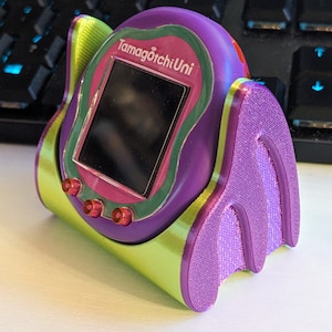May include: A purple and green Tamagotchi Uni device with a black screen and three red buttons. The device is in a purple and green holder. The text "Tamagotchi Uni" is visible on the screen.