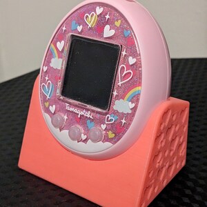 May include: A pink Tamagotchi virtual pet with a decorative face featuring hearts, rainbows, and the word "Tamagotchi". It rests in a coral-coloured stand. The device has three pink buttons below a black screen.