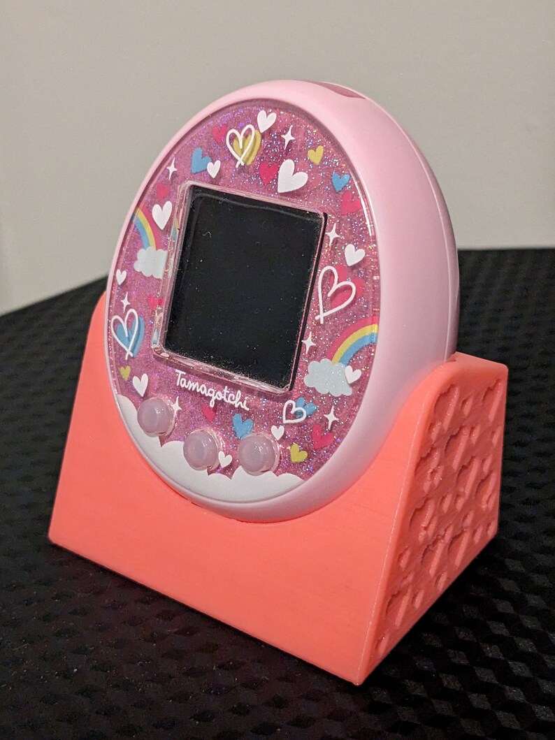 May include: A pink Tamagotchi virtual pet with a decorative face featuring hearts, rainbows, and the word "Tamagotchi". It rests in a coral-coloured stand. The device has three pink buttons below a black screen.