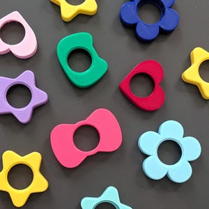 May include: A collection of brightly coloured, 3D-printed shapes, including stars, flowers, hearts, and bowties. The shapes are in yellow, pink, green, red, blue, and purple. Each shape has a circular hole in the centre.