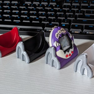 Puede incluir: Cuatro soportes coloridos impresos en 3D para un pequeño dispositivo electrónico. Los soportes son rojos, negros, blancos y un dispositivo morado y blanco con el texto "DEVIL GOTCHI". Tienen un diseño de garra con detalles plateados.