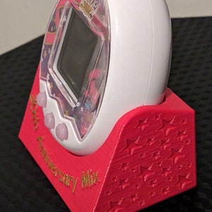 May include: A white digital pet device with a clear screen, in a red stand. The device has a colourful design with the text "20th Anniversary Mix". The red stand is decorated with star patterns.