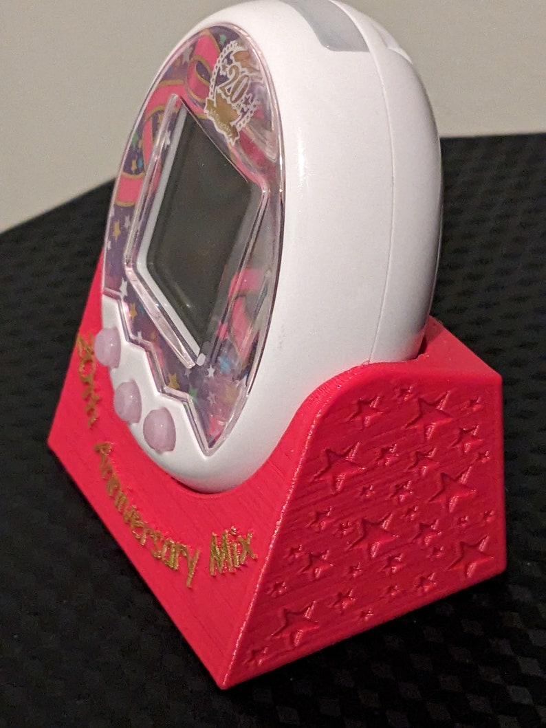 May include: A white digital pet device with a clear screen, in a red stand. The device has a colourful design with the text "20th Anniversary Mix". The red stand is decorated with star patterns.