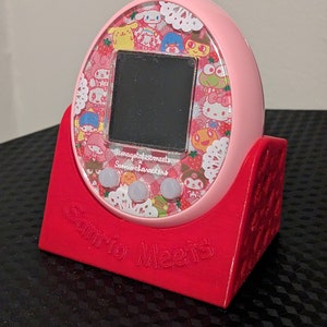 May include: A pink Tamagotchi device with a screen and buttons, on a red stand. The device has a colourful design with Sanrio characters and the text "Sanrio Meets".