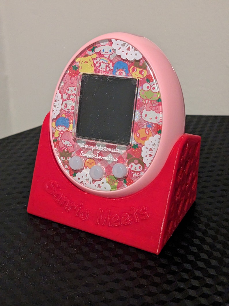 May include: A pink Tamagotchi device with a screen and buttons, on a red stand. The device has a colourful design with Sanrio characters and the text "Sanrio Meets".