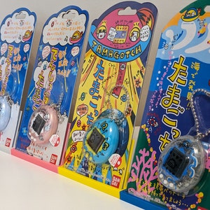 Puede incluir: Cuatro juguetes virtuales Tamagotchi en su embalaje original, cada uno con un esquema de color diferente. El embalaje presenta texto e ilustraciones japonesas. Los Tamagotchi son de color azul, rosa y turquesa, con una pequeña pantalla y un llavero.