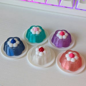 Puede incluir: Cinco gelatinas coloridas con forma de flor sobre platillos blancos. Las gelatinas son de color azul, turquesa, morado, transparente y rosa, cada una coronada con un centro blanco y rojo o rosa contrastante. El fondo incluye un escritorio blanco y un teclado.