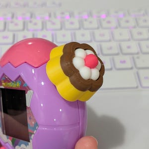 May include: A handheld, purple and pink electronic toy with a decorative, cupcake-shaped attachment. The attachment features yellow, brown, white, and pink elements. The toy has a small screen and the word "Tamagotchi" is visible.