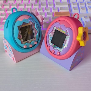 以下が含まれることがあります： パステルカラーの2つのTamagotchiバーチャルペットデバイス。1つはティールとピンク、もう1つはピンクと紫で、それぞれ小さな画面にピクセル化された画像が表示されます。デバイスは、パステル模様と「Tamagotchi」の文字が入った装飾的なスタンドに置かれています。