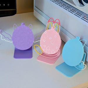 May include: Three colorful bookends in purple, pink, and blue. Each bookend features a rounded shape with decorative elements: wings, hearts, and a curved design. The bookends are made of a solid material and stand upright on a flat base.