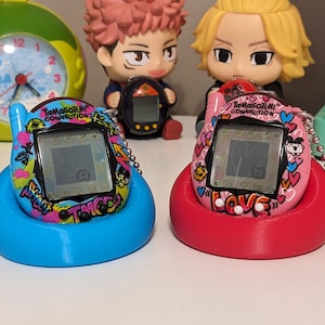 May include: Two Tamagotchi Connection digital pets, one blue and one pink, sit in matching stands. The blue one has a graffiti-style design, while the pink one is decorated with hearts and the word "LOVE". Both have small screens.