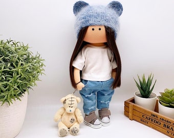 Une poupée-cadeau faite à la main vêtue d'un t-shirt blanc, d'un chapeau avec de petites oreilles et d'un jean déchiré, accompagnée d'un petit ours en peluche