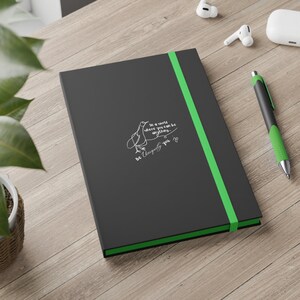 Op de afbeelding: Een zwart notitieboek met een groene elastische bandsluiting en groene rand. Het notitieboek bevat witte tekst en een vogelillustratie met de tekst "In a world where you can be anything... Be uniquely you."